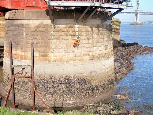 Image of maintenance worker inspecting the base of one of the towers of the Forth Bridge near the waterline